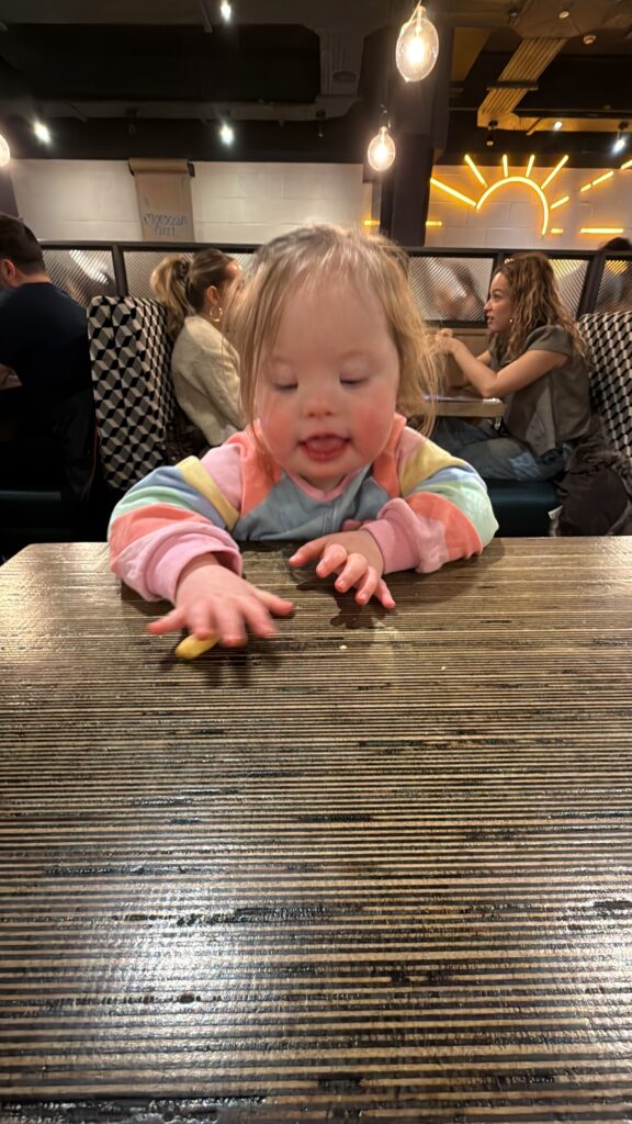 Young girl with Down syndrome sitting at a wooden restaurant table, reaching forward with her hand to pick up a small piece of food while concentrating on the movement. Warm pendant lights glow above and other diners sit in the background, capturing an everyday moment of growing independence and highlighting the steady progress seen in down syndrome development.