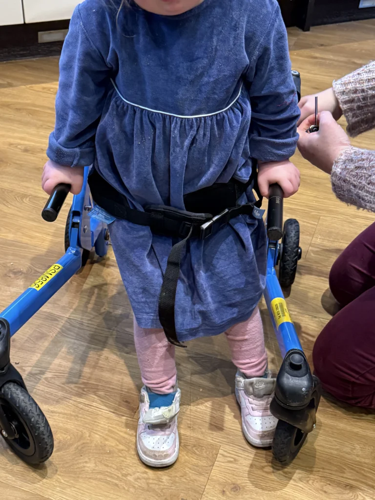 Close-up of a young girl with Down syndrome standing inside a blue walking frame, gripping the black handles while a caregiver adjusts the support strap around her waist. Her feet are planted on a wooden floor as she practices balance and weight bearing, illustrating an important stage in down syndrome development and growing physical independence.
