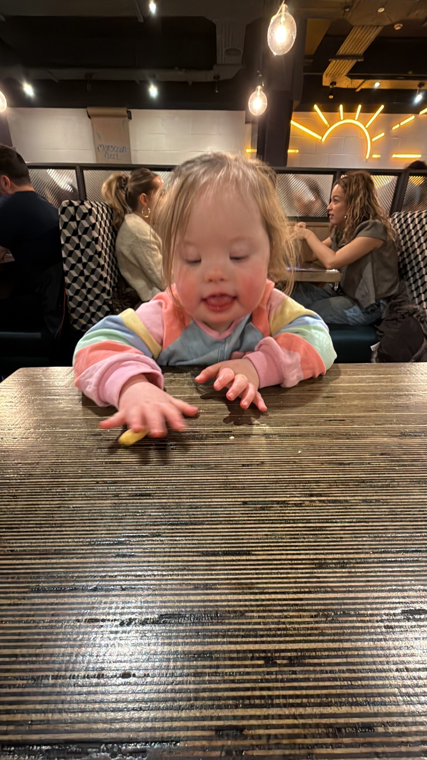 Young girl with Down syndrome sitting at a wooden restaurant table, reaching forward with her hand to pick up a small piece of food while concentrating on the movement. Warm pendant lights glow above and other diners sit in the background, capturing an everyday moment of growing independence and highlighting the steady progress seen in down syndrome development.