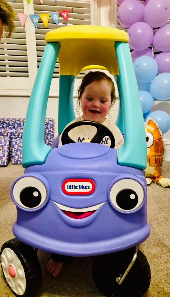 Smiling young girl with Down syndrome sitting in a purple Little Tikes toy car decorated with birthday balloons in the background. She grips the steering wheel proudly, celebrating independence, play skills and joyful progress in her down syndrome development journey.