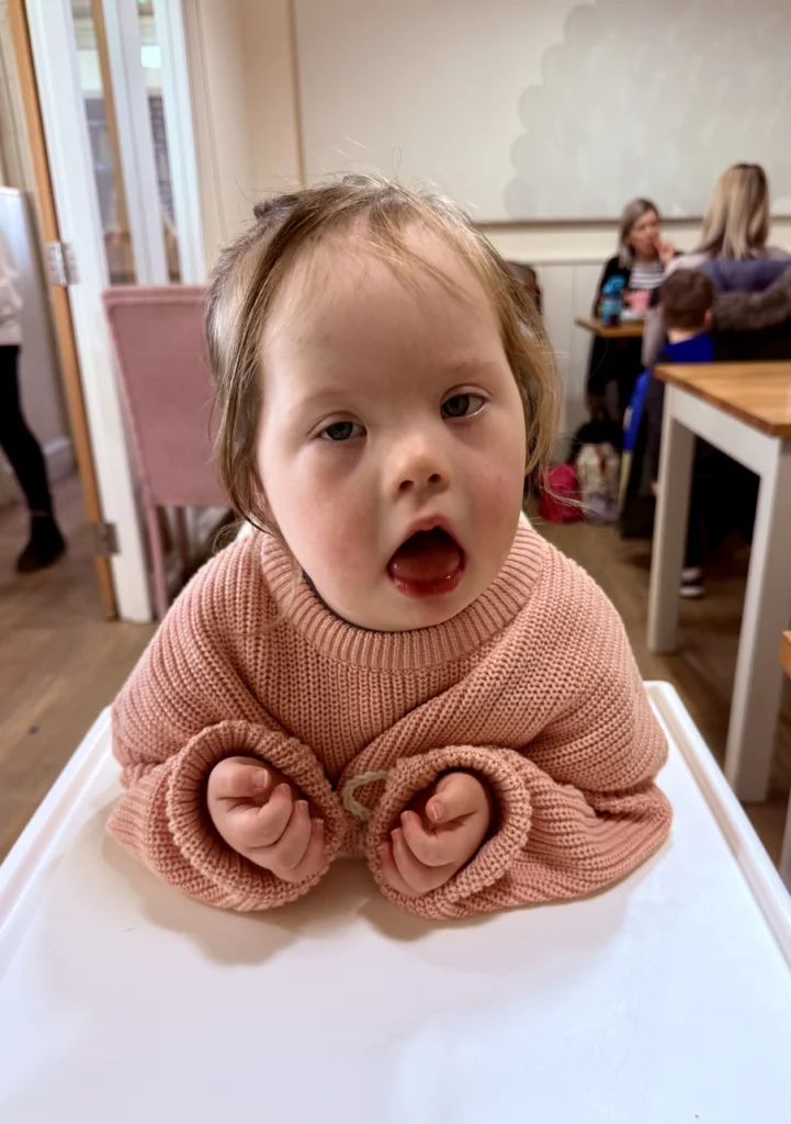 Close-up of a young girl with Down syndrome sitting in a high chair at a café, wearing a soft pink knitted jumper and looking forward with her mouth slightly open. Her small hands rest on the tray in front of her while other diners sit in the background, capturing a quiet everyday moment that reflects emotional connection and steady progress in down syndrome development.
