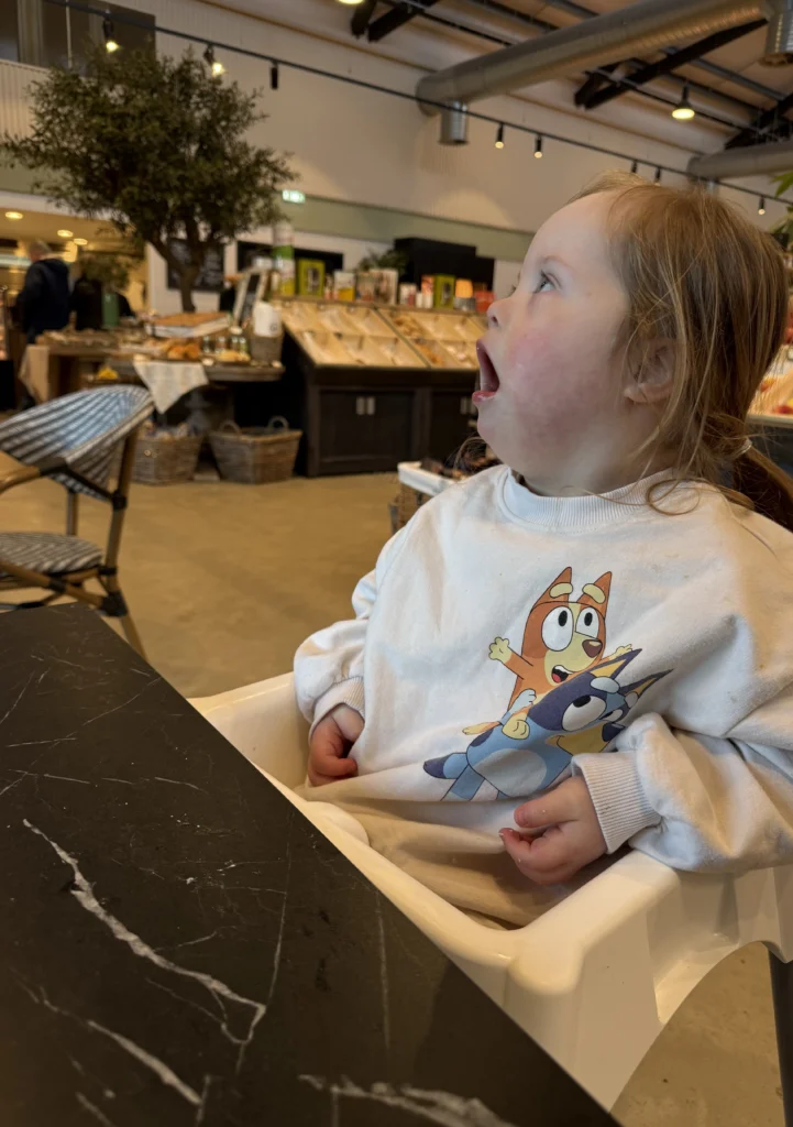Young girl with Down syndrome sitting in a high chair at a café, wearing a sweatshirt with cartoon characters and looking upward with her mouth slightly open in curiosity. The café interior with wooden displays and warm lighting surrounds her, capturing a moment of awareness and engagement that reflects cognitive and social progress in down syndrome development.