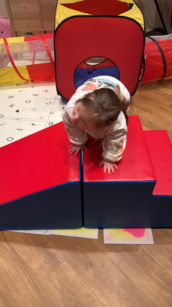 Young girl with Down syndrome crawling on her hands and knees across red and blue foam play blocks positioned in front of a colourful play tunnel. Her palms press firmly into the padded surface as she shifts her weight forward, demonstrating coordination, balance and an important gross motor stage in down syndrome development.