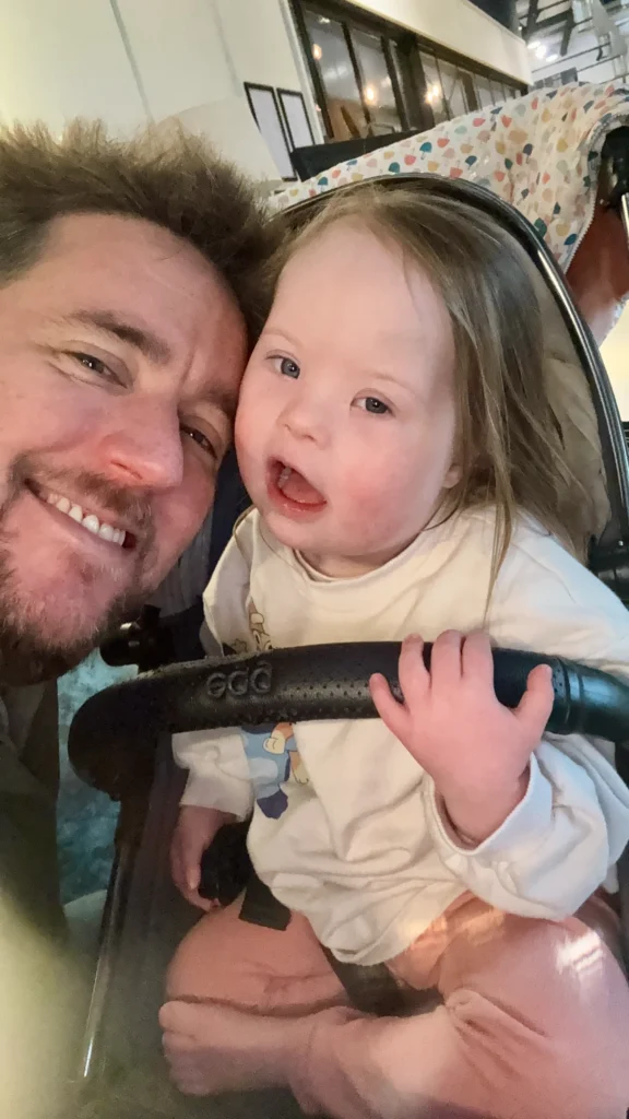Smiling father leaning close to his young daughter with Down syndrome as she sits safely in a stroller, her small hand resting on the front bar. They look toward the camera together, capturing connection, security and the social-emotional growth that plays an important role in down syndrome development.