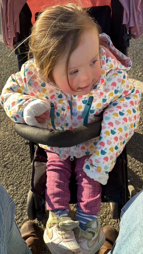 Young girl with Down syndrome sitting in a stroller outdoors on a sunny day, wearing a colourful umbrella-patterned coat and pink leggings. She leans slightly forward and looks down with curiosity, showing independence and engagement with her surroundings as part of her ongoing down syndrome development.