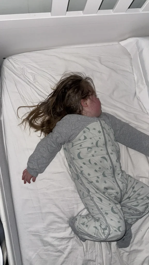 Young girl with Down syndrome sleeping peacefully in her cot, lying on her side with one arm stretched outward and wearing a grey long-sleeved top and star-patterned sleep suit. The calm bedtime setting captures the importance of rest and routine in supporting healthy down syndrome development and overall wellbeing.