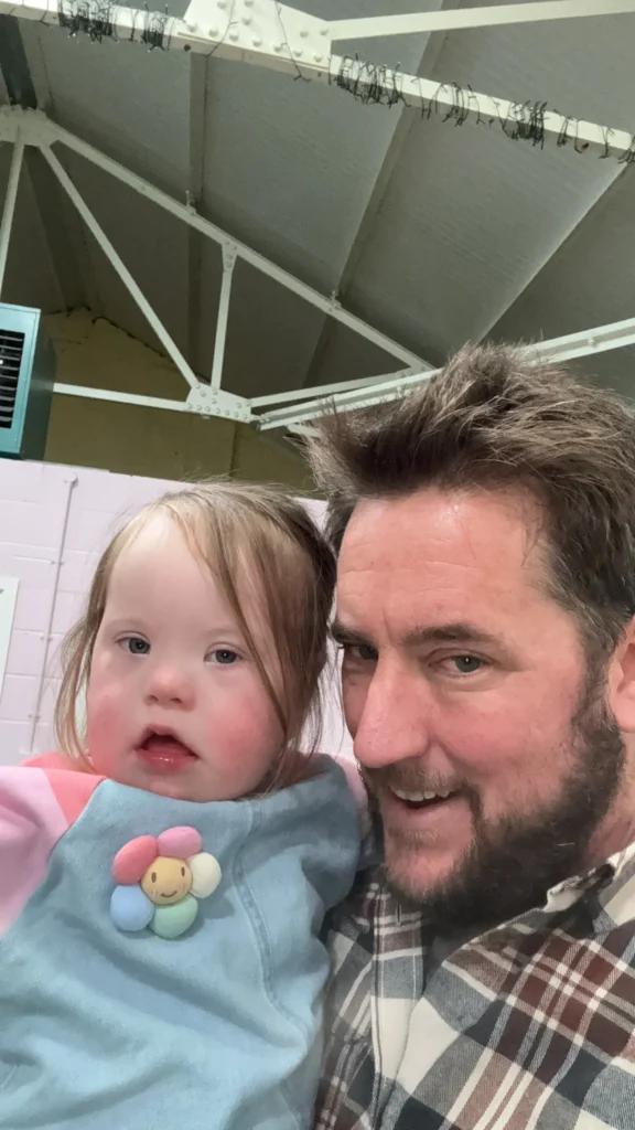 What My Daughter’s Fourth Birthday Taught Me About Down Syndrome Milestones 12 Close-up selfie of a father holding his young daughter with Down syndrome inside a large indoor space with exposed beams overhead. She rests comfortably in his arms wearing a pastel sweatshirt, while he smiles beside her, capturing a quiet, supportive moment that reflects the love behind every developmental milestone and the family journey through down syndrome milestones experienced by children with Down syndrome.