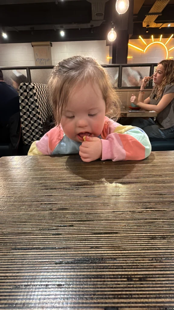 What My Daughter’s Fourth Birthday Taught Me About Down Syndrome Milestones 6 Young girl with Down syndrome sitting at a wooden restaurant table, bringing a small piece of food to her mouth with her fingers while concentrating on eating independently. Warm hanging lights glow above and other diners sit in the background, capturing an everyday developmental milestone in her down syndrome milestones journey and highlighting the growing independence of children with Down syndrome.