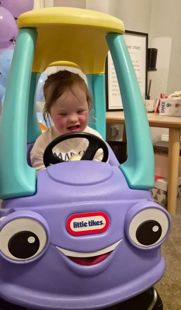What My Daughter’s Fourth Birthday Taught Me About Down Syndrome Milestones 4 Four-year-old girl with Down syndrome sitting inside a purple and blue Little Tikes toy car, smiling as she grips the black steering wheel during her birthday celebration. Purple and blue balloons and a “Happy Birthday” banner hang behind her, marking an important moment in her down syndrome milestones and celebrating each developmental milestone reached by children with Down syndrome.