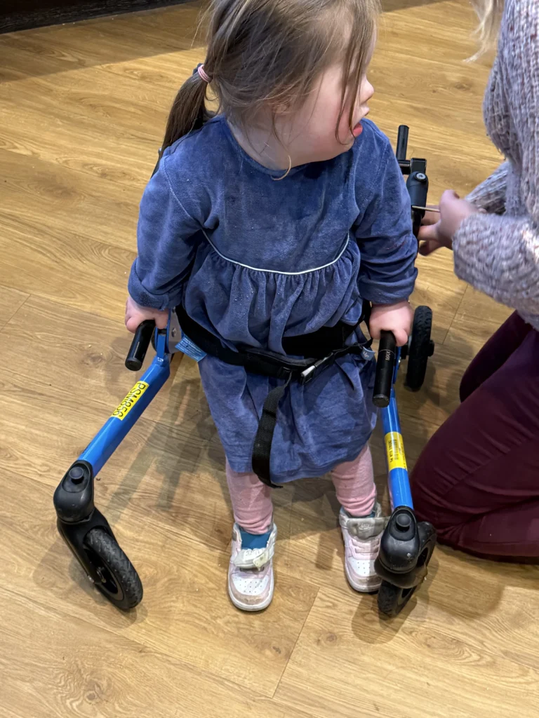 What My Daughter’s Fourth Birthday Taught Me About Down Syndrome Milestones 7 Four-year-old girl with Down syndrome standing on a wooden floor using a small blue walking frame, gripping the black handles while an adult kneels beside her adjusting the support strap at her waist. She looks to the side as she balances on her own two feet, representing an important developmental milestone in her journey of down syndrome milestones and highlighting the steady progress children with Down syndrome make with mobility and independence.