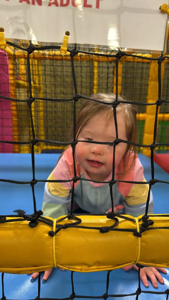Young girl with Down syndrome crawling forward inside a soft play area while looking through black safety netting and supporting her weight on a blue padded mat. This moment highlights Down syndrome hypotonia and how low muscle tone in Down syndrome can influence early gross motor development and balance.