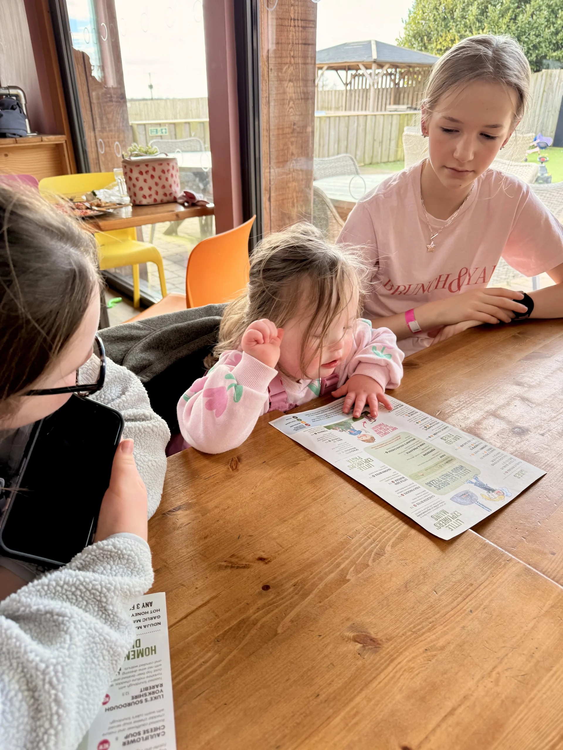 Young child with Down syndrome sitting at a table with older siblings while looking at a leaflet, illustrating down syndrome communication strategies that support down syndrome language development and early interaction.