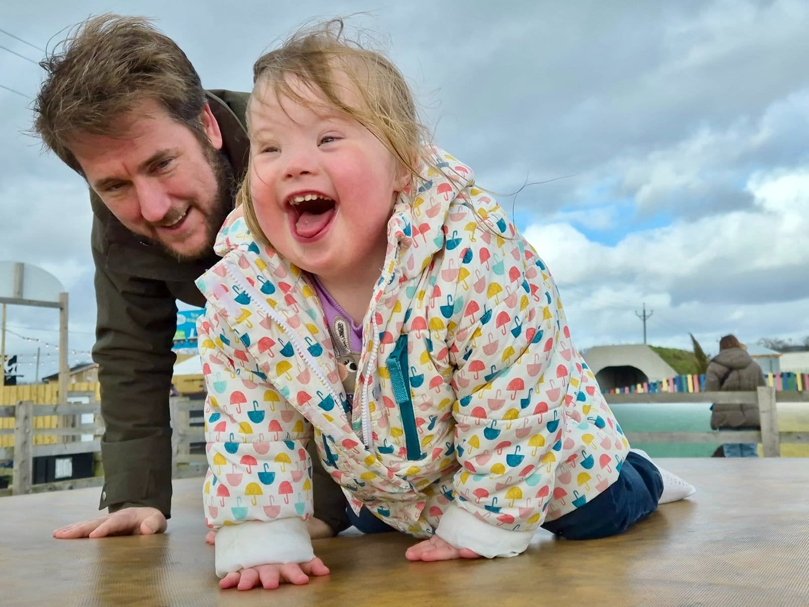 Father playing with his young daughter with Down syndrome as she laughs while crawling on a playground platform, illustrating joyful interaction that supports language and communication development in Down syndrome, including down syndrome speech and language growth through everyday play and connection.