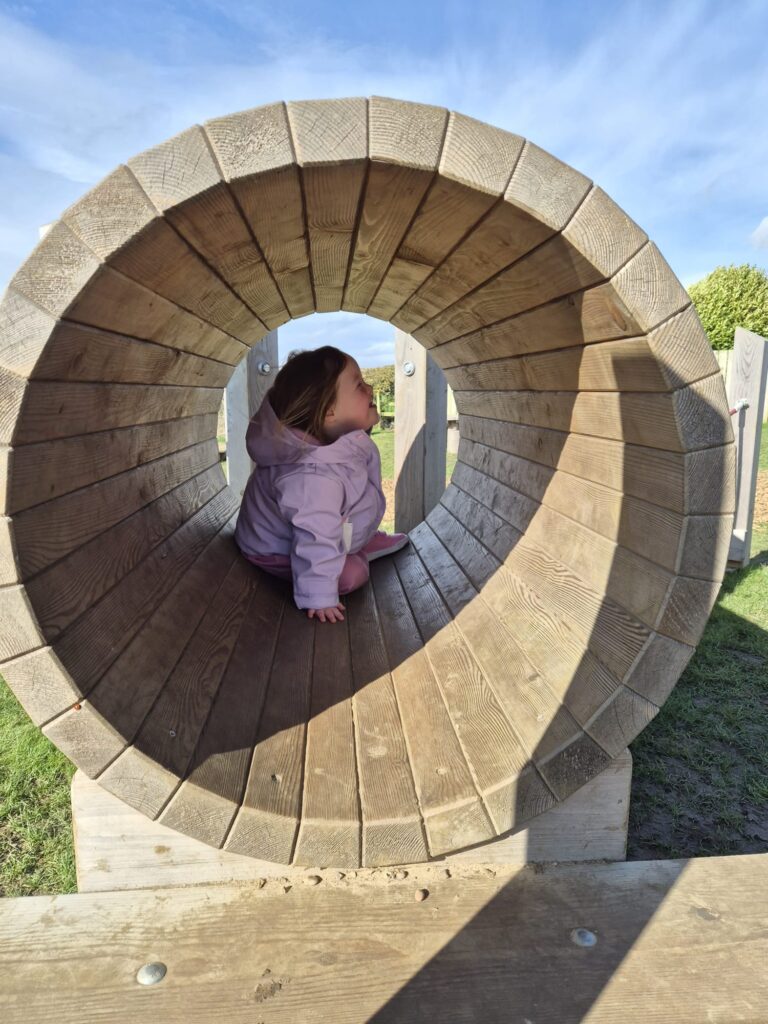 Toddler with Down syndrome sitting inside a wooden playground tunnel while exploring her surroundings, reflecting playful environments that support language and communication development in down syndrome and early social interaction.