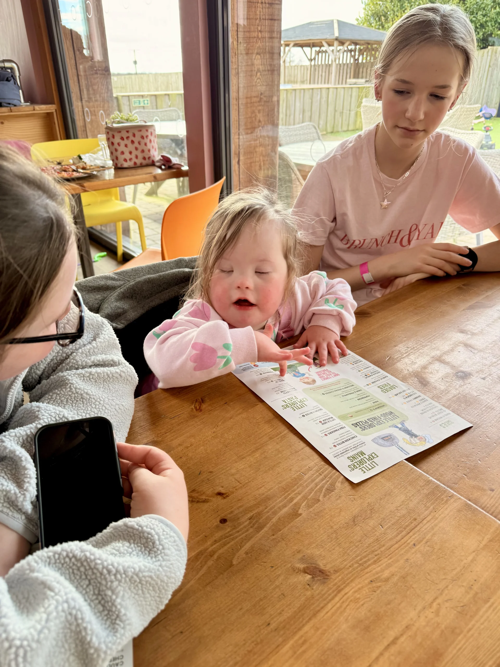 Young child with Down syndrome pointing at a leaflet while sitting with older children at a table, showing shared attention that can help address speech delay in children with down syndrome and support down syndrome speech and language development.