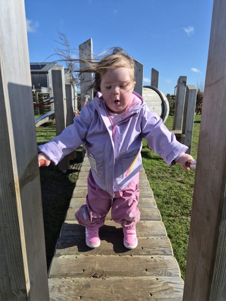 Child with Down syndrome walking across a wooden playground balance bridge while concentrating, highlighting confidence and independence often seen alongside speech characteristics of down syndrome and ongoing down syndrome and speech development.