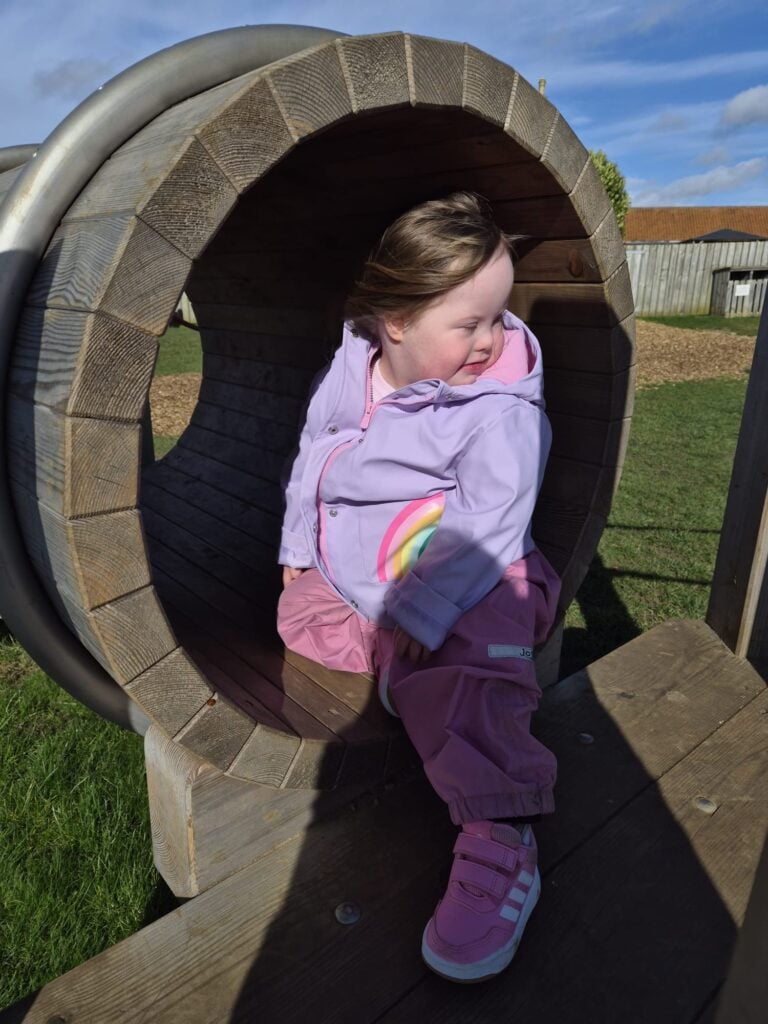 Young child with Down syndrome sitting inside a wooden playground tunnel and looking back with a smile, representing everyday play that encourages speech and language difficulties to be addressed through interaction and supports speech delay down syndrome progress.
