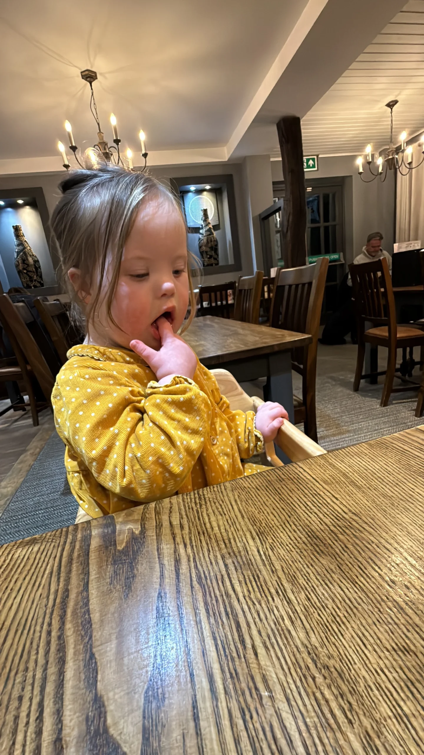 Young child with Down syndrome sitting in a restaurant high chair and exploring her mouth with her fingers, illustrating common speech characteristics of Down syndrome and early stages of down syndrome speech and language development.